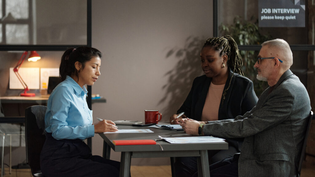 A woman and a man interviewing another woman at a table with paperwork and a coffee cup on the table.